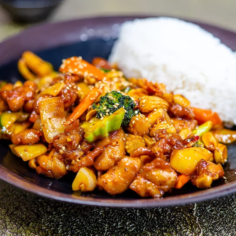 Teriyaki chicken with broccoli, carrots, and bell peppers, served with white rice on a dark plate.